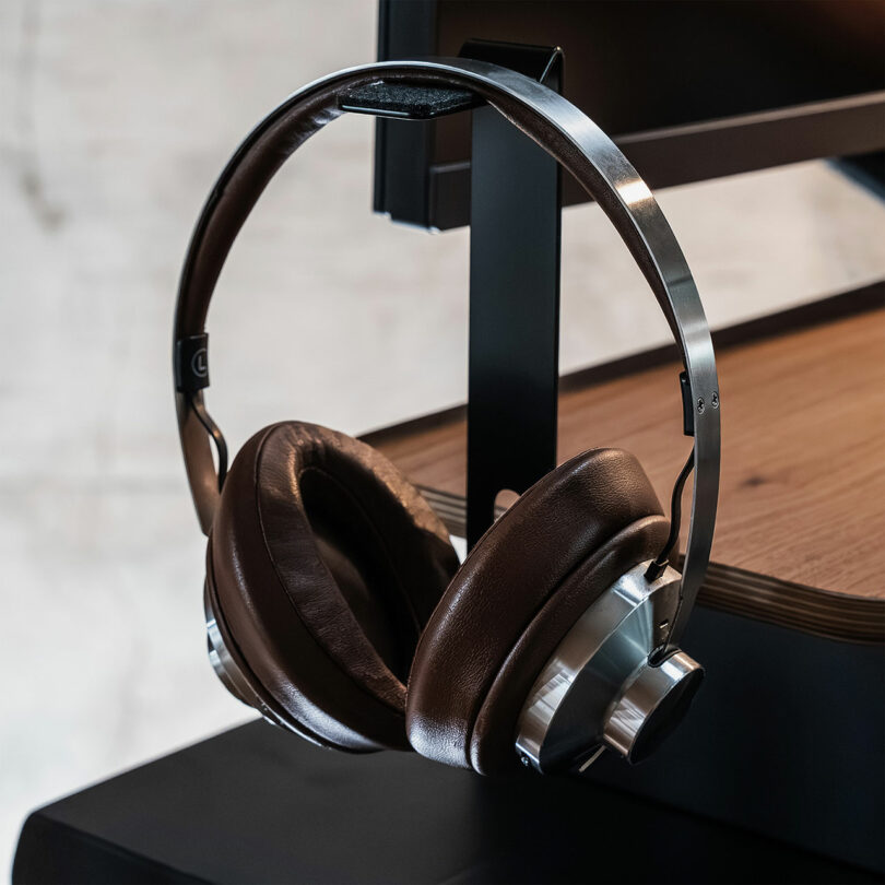 A pair of headphones hanging on a black metal Balolo stand attached to the side of a wooden desk.