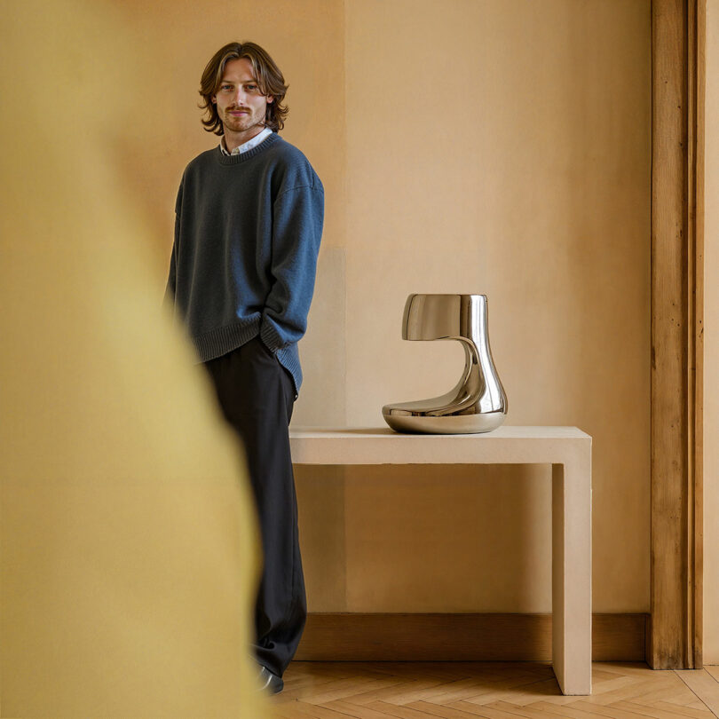 Paddy Pike standing next to a metallic sculptural object in a minimalist setting.