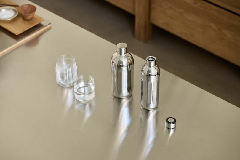Two Aarke To-Go Bottles of different sizes next to two glasses of sparkling water on a metallic surface.