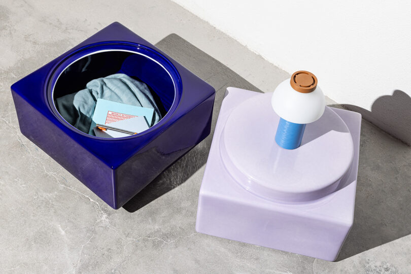 Two ceramic containers on a floor; one is open containing fabric and paper, the other is closed serving as a base for a lamp.