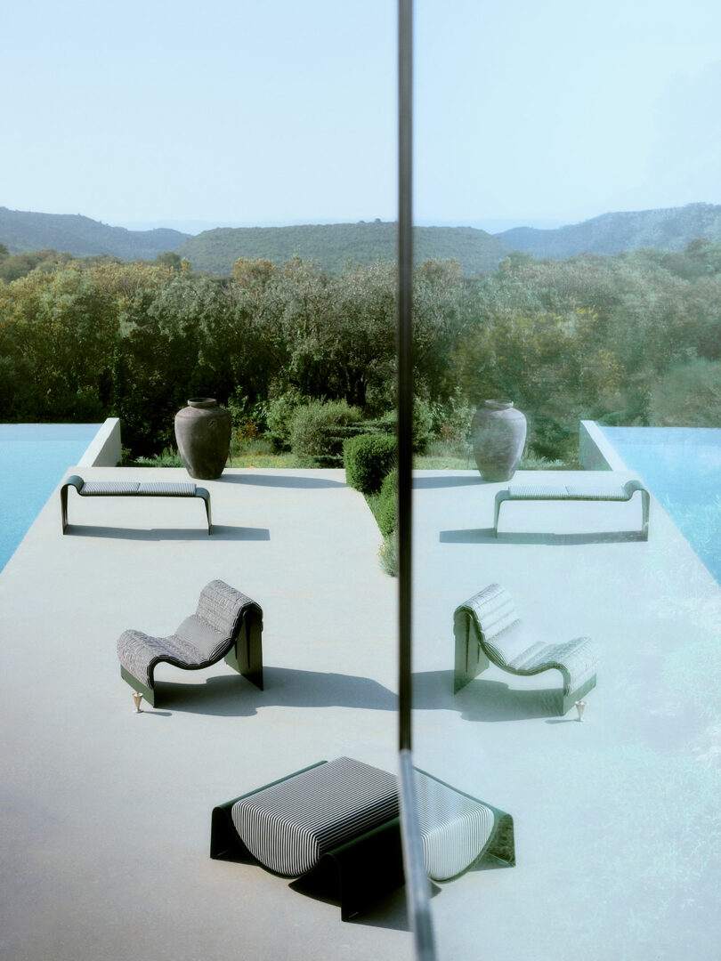 Modern poolside patio with lounge chairs reflected in a glass wall against a hilly natural landscape.
