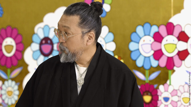 Takashi Murakami in a black robe standing before a gold floral mural.