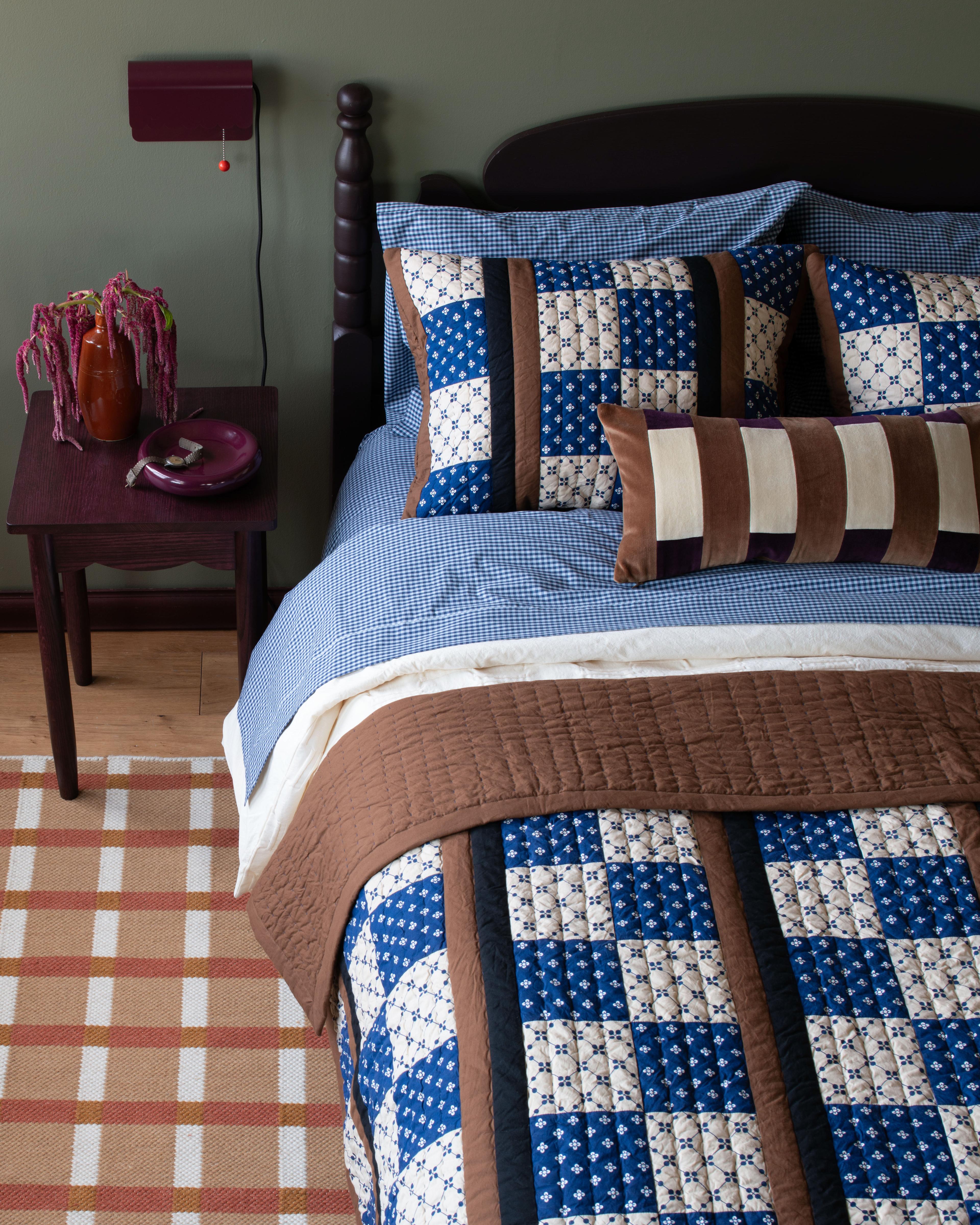 A bed styled with a mix of a thick comforter, a quilt, and a rug placed underneath.