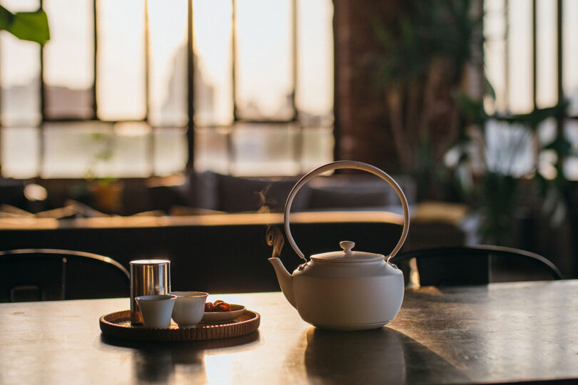 A white teapot on a table with warm sunlight streaming through a large window.