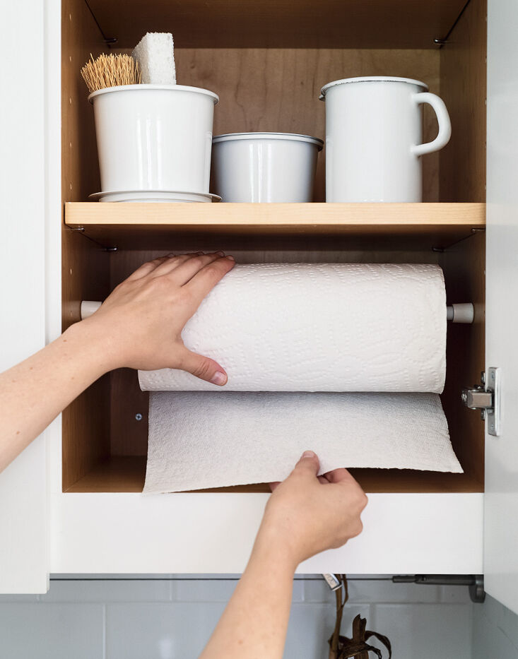 A white tension rod installed horizontally between two cabinets holding a paper towel roll.