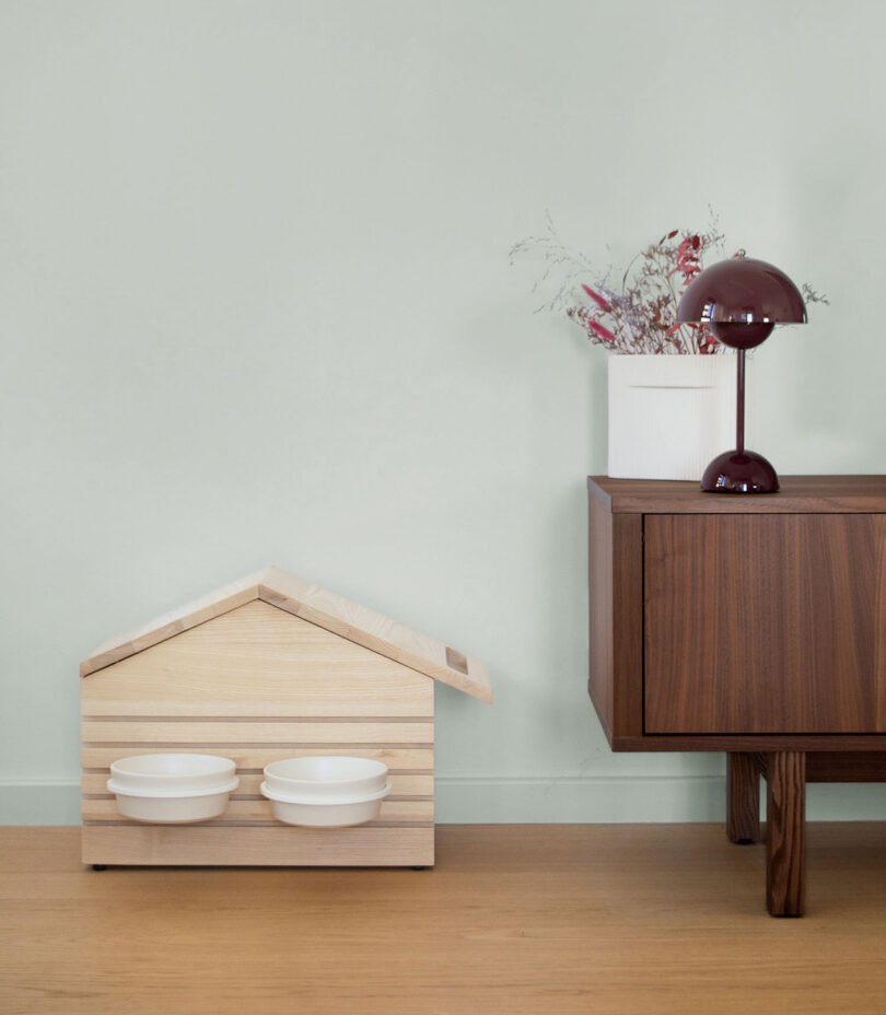 A house-shaped wooden pet feeder with two bowls placed on a light wood floor next to a red lamp.