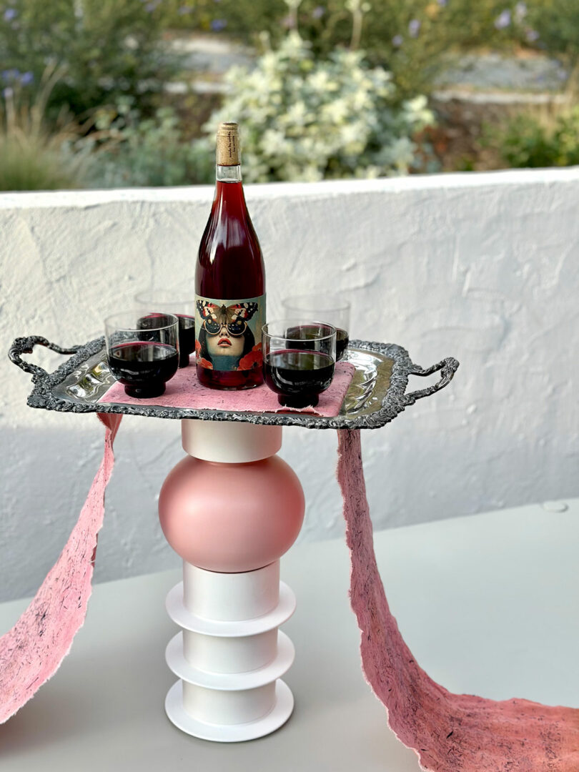 An ornate silver tray with wine bottles and glasses staged on a modern pink-draped sculptural pedestal.