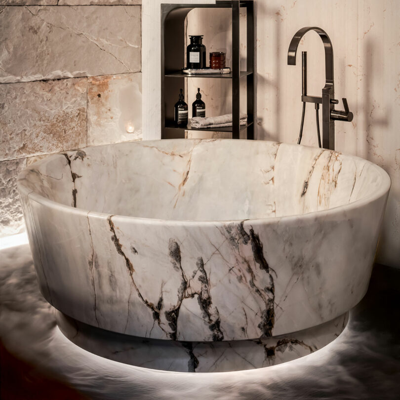 A round, solid-carved marble bathtub with intricate gray and brown veining in a modern stone bathroom.