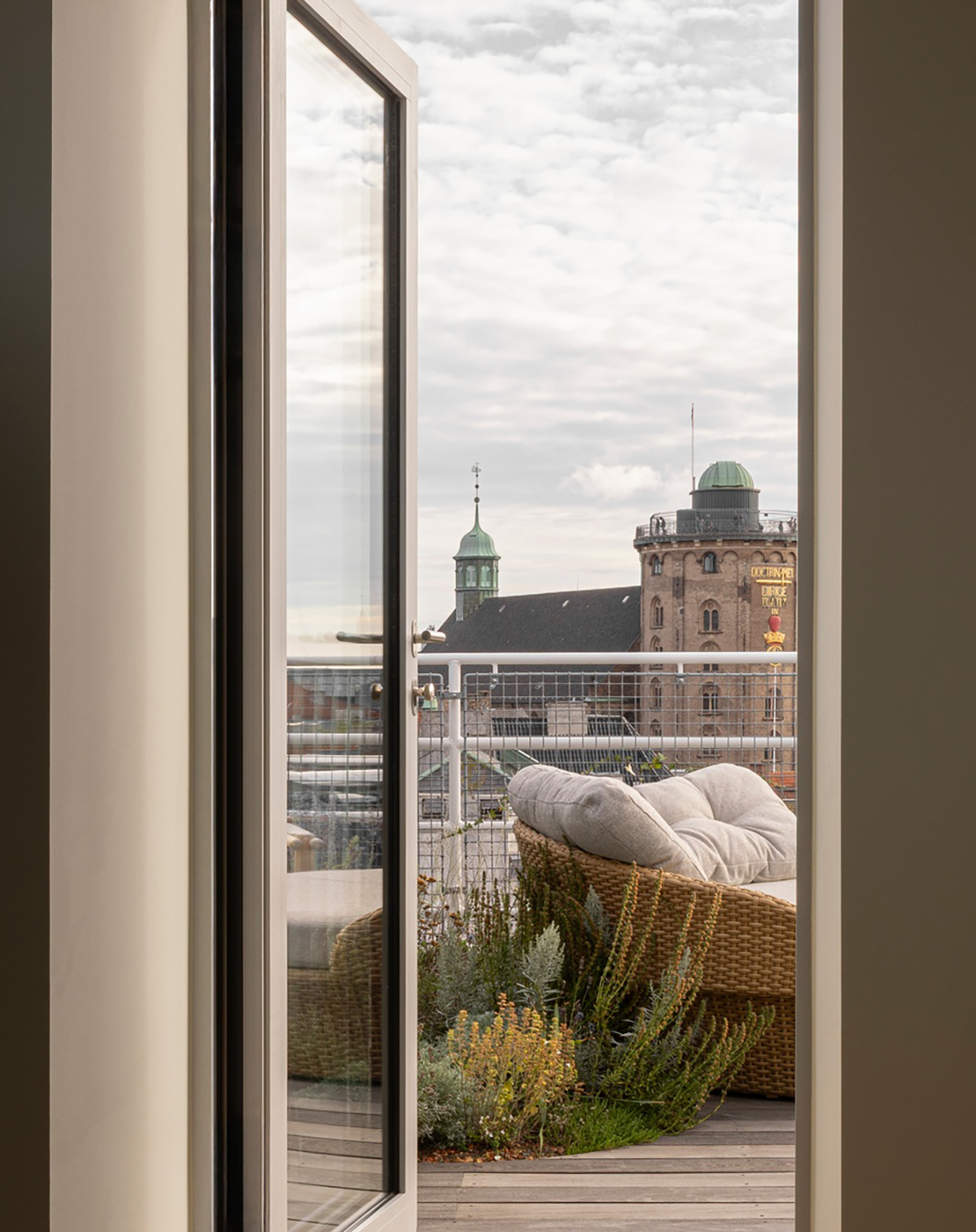 View from a hotel balcony with a wicker chair, looking out over Copenhagen city rooftops and green domes.