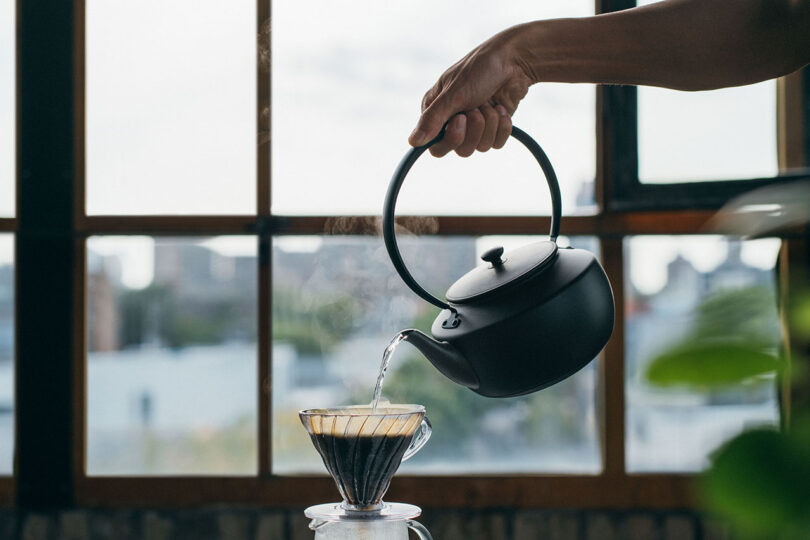 A hand pouring hot water from a black kettle into a glass coffee dripper by a window.
