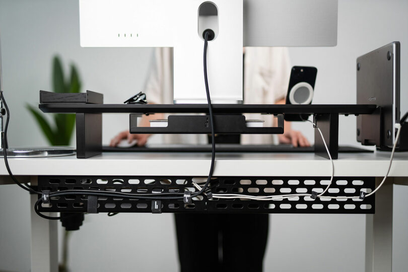 A person standing at a desk with a Balolo monitor stand and a visible cable management tray underneath.