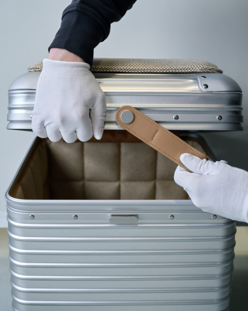 Close-up of gloved hands opening the aluminum stool to reveal the tan leather interior strap and lining.