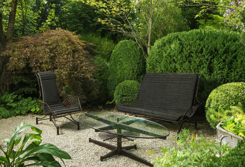 A set of metal patio furniture including a bench and glass-top table in a lush, gravel-floored garden.