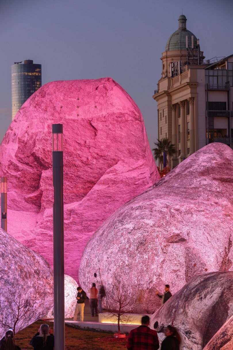 Large boulders glowing with pink light in a city square at dusk with skyscrapers in the background.