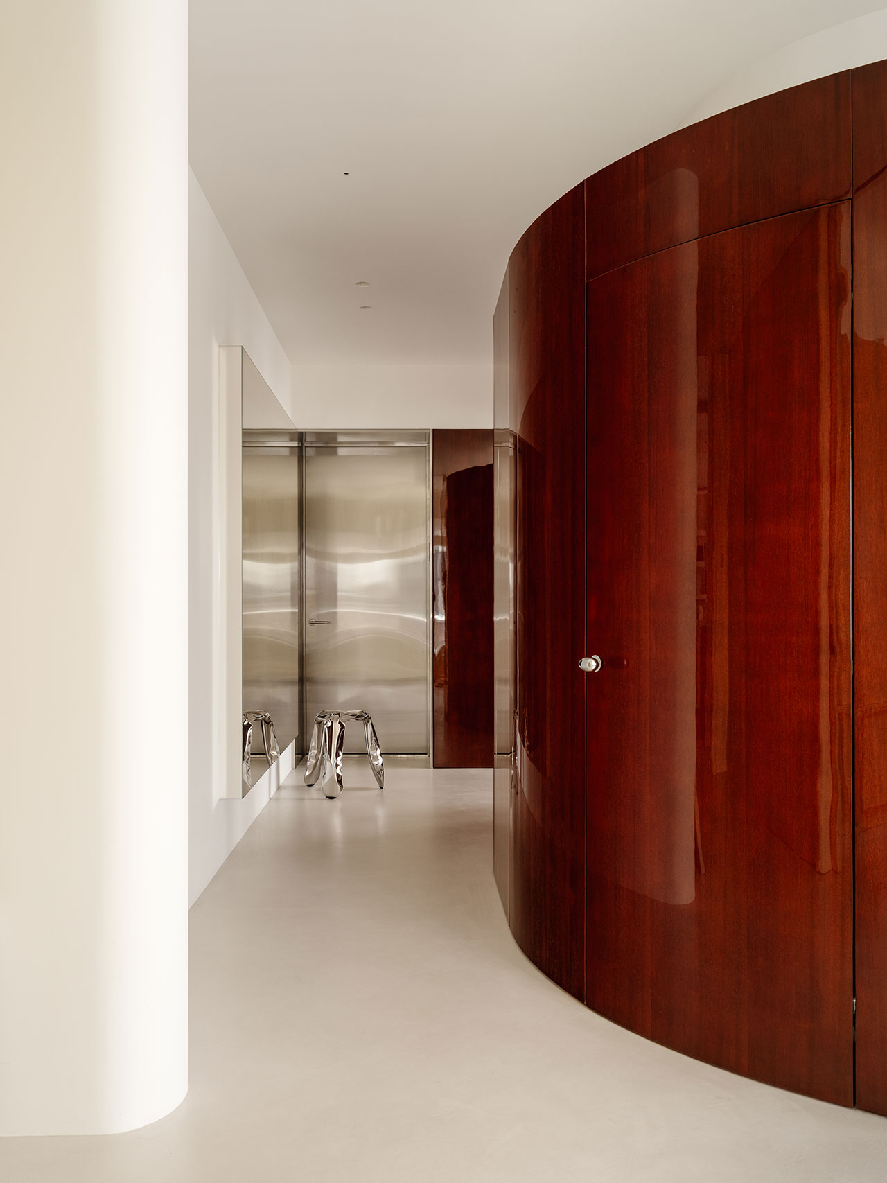 Minimalist hallway with polished wood curved wall, stainless steel door, and reflective stool.