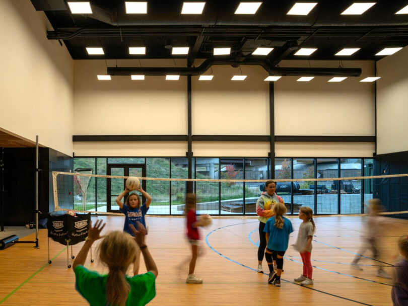 Children playing volleyball in a large indoor court with polished wood floors and high ceilings within the campus.