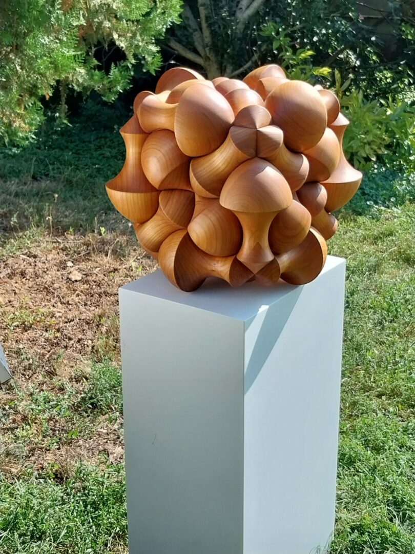 A large geometric wooden sculpture based on the sphericon principle on a white pedestal outdoors.