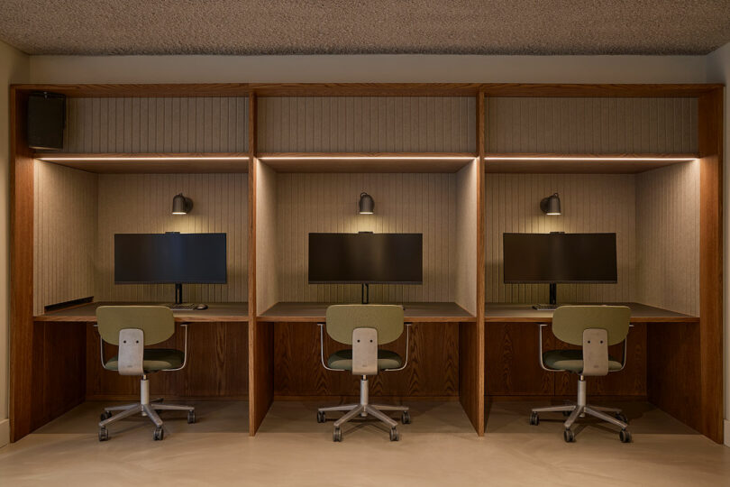 Three minimalist wooden office desks with monitors and individual lamps against a neutral wall.