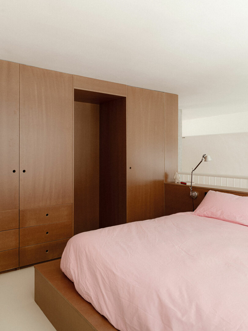 A minimalist bedroom with built-in wooden wardrobes and a pink duvet.