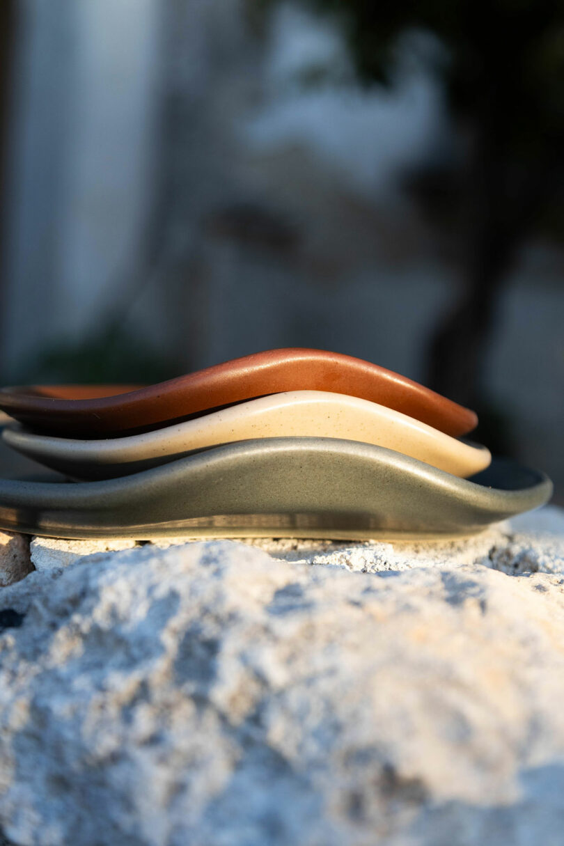 Three wavy ceramic plates stacked on a rough stone surface with a soft outdoor background.