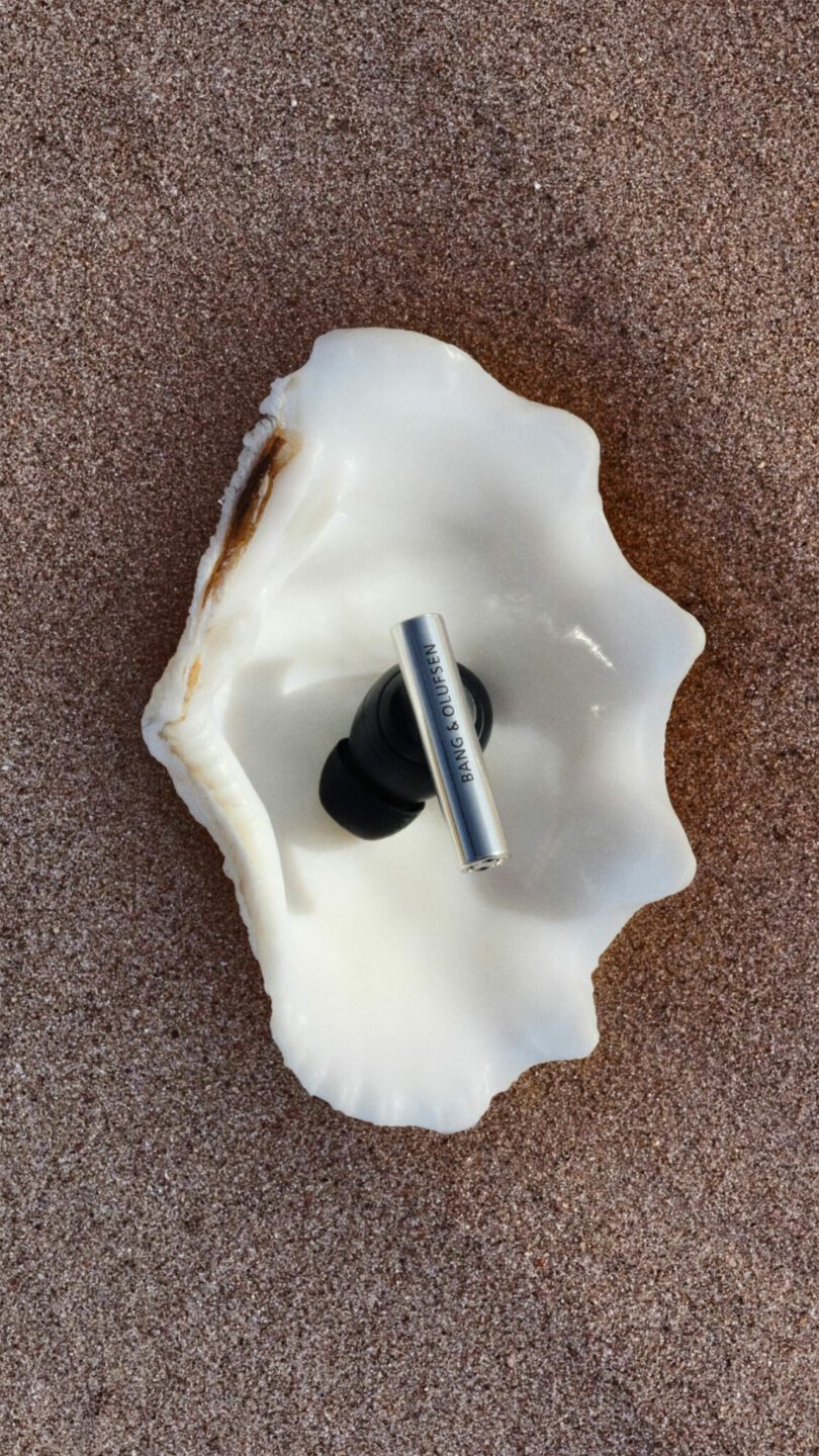 A metallic wireless earbud placed inside a white seashell on a sandy surface.