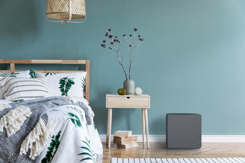 A gray air purifier on the floor of a bedroom with a blue-green wall.
