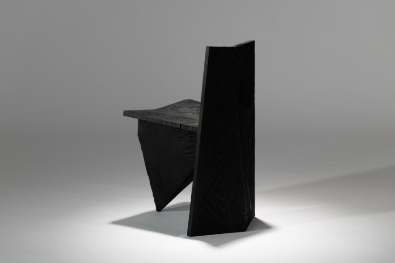 A black wooden chair with sharp geometric lines and a minimalist profile against a gray background.