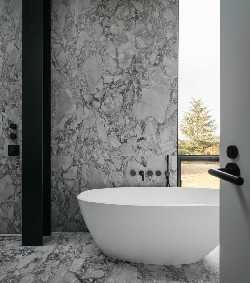Modern bathroom with gray marble walls and a freestanding white bathtub facing a window.