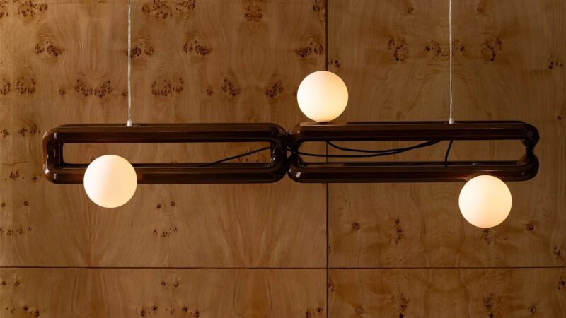 A modern three-bulb chandelier against a warm wood-paneled wall.