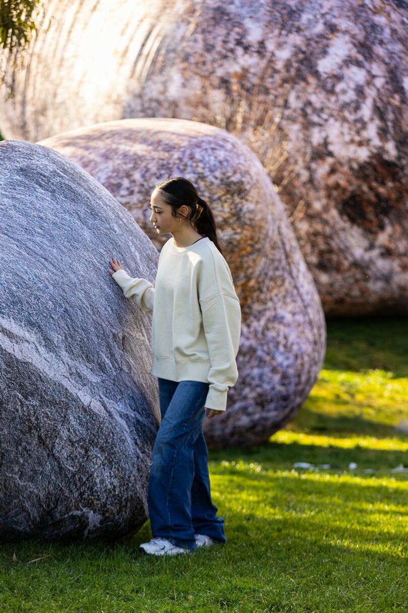 A person touching a large inflatable rock resting gently on green grass.