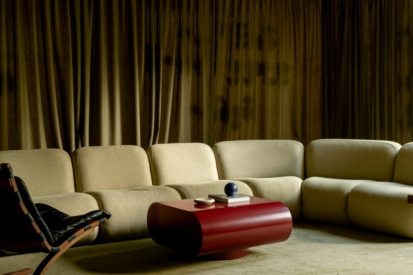 A modern living area featuring a beige curved sectional sofa, a red oval coffee table, and heavy brown curtains.