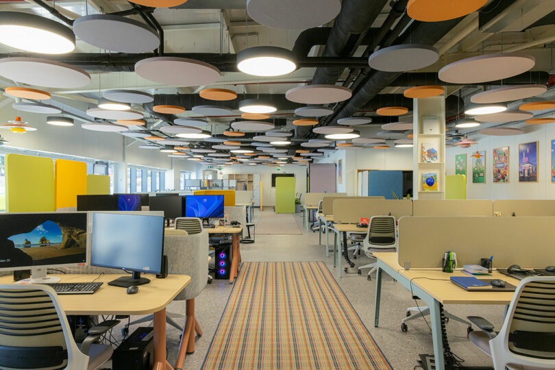 A wide shot of a modern open-plan office with ergonomic chairs, computers, and colorful ceiling acoustics.