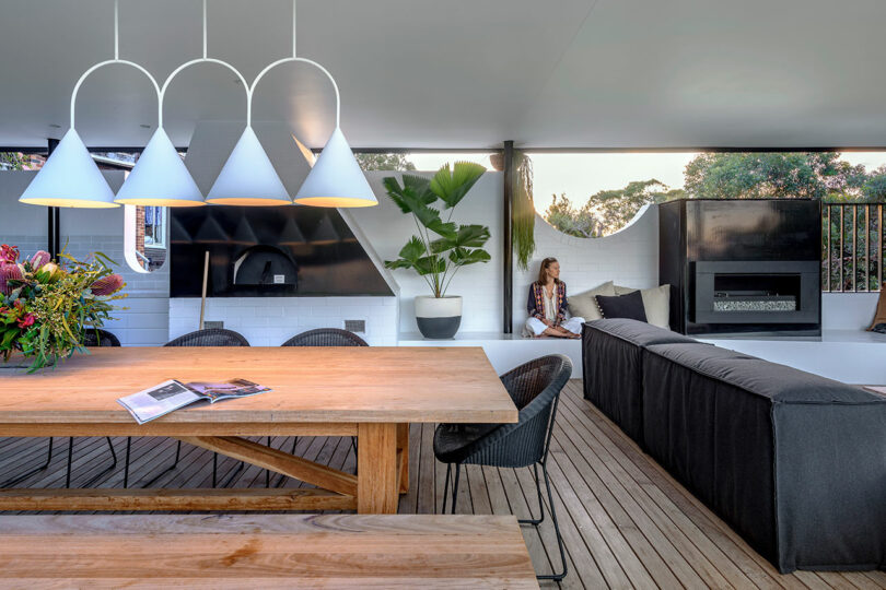 A woman lounging on a black sectional sofa in a covered outdoor living room with a fireplace and modern pendant lighting.