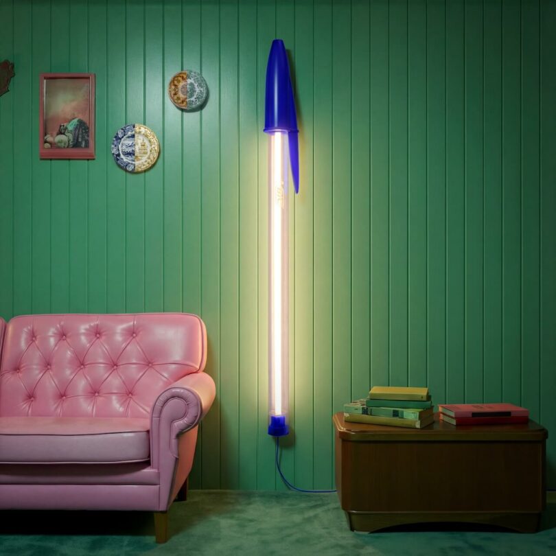 A blue Seletti BIC floor lamp shaped like a giant pen standing next to a pink sofa.