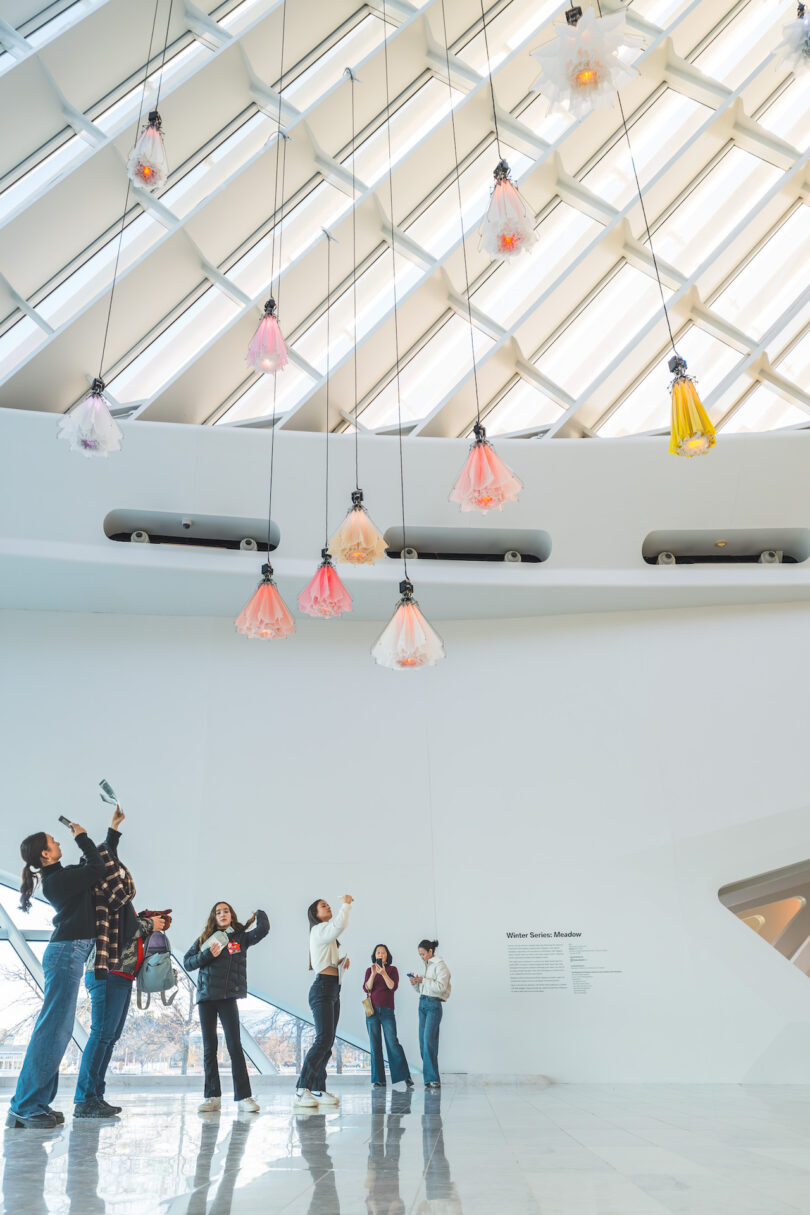 Visitors standing in a white gallery space looking up at suspended kinetic floral sculptures.