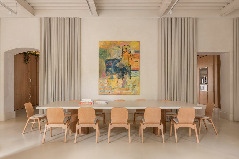 A long wooden communal table with ten chairs in a neutral-toned room with arched entryways.