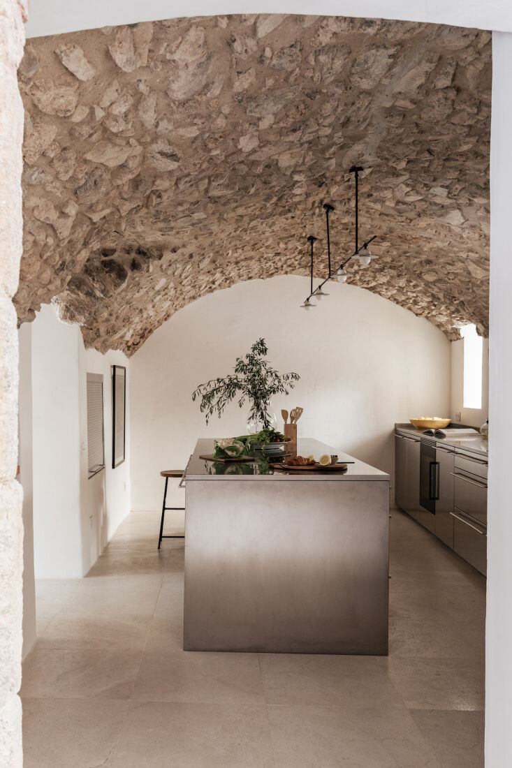 A modern stainless steel kitchen island set against a backdrop of ancient vaulted stone ceilings.