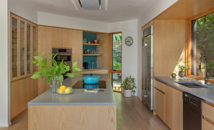 Bright modern kitchen with light wood cabinetry, center island, and large windows overlooking trees.