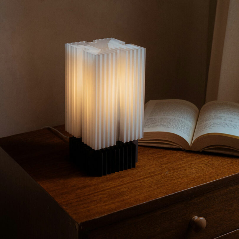 A modern ribbed desk lamp emitting warm light on a wooden surface next to an open book.