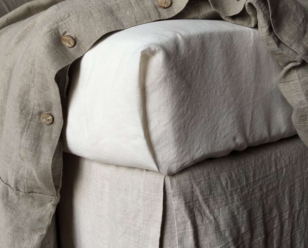 Hands tucking a large flat linen sheet tightly around the corner of a mattress.