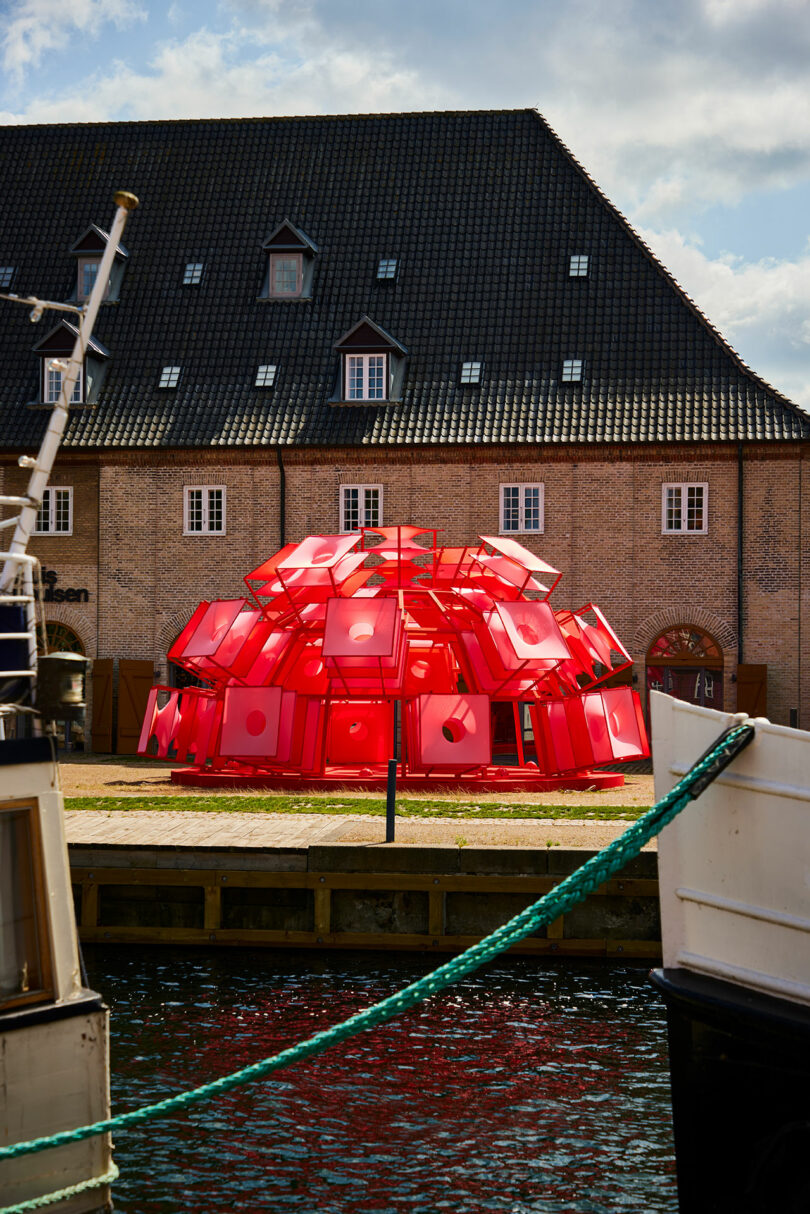 The red dome-shaped installation situated on a lawn in front of a traditional brick building by a canal.