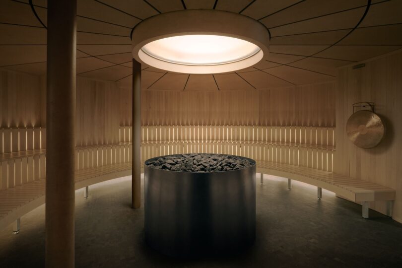 Large circular wooden sauna with tiered seating and a central stone heater under a skylight.