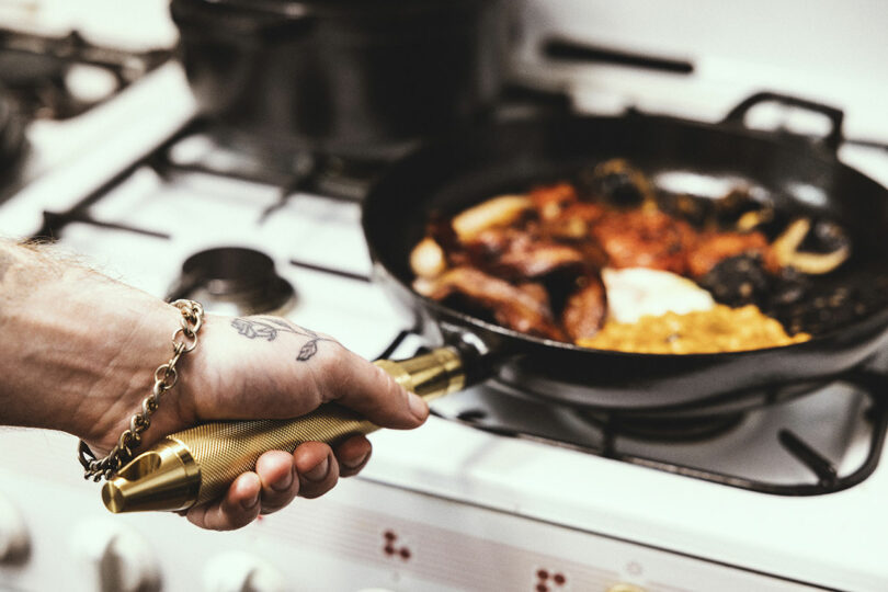 A person searing food in a black Staub frying pan on a professional stove, holding the solid brass handle.