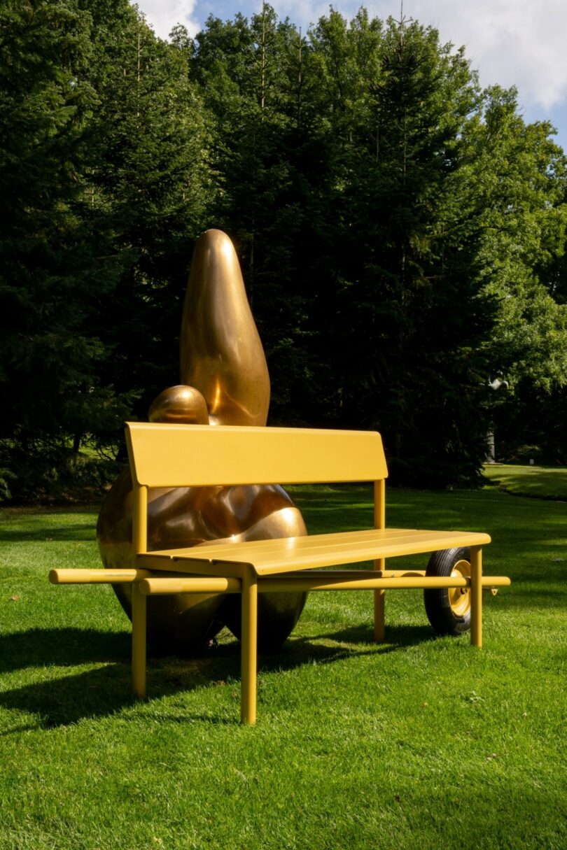 A yellow Wheelbench positioned on a lawn in front of a large bronze abstract sculpture.
