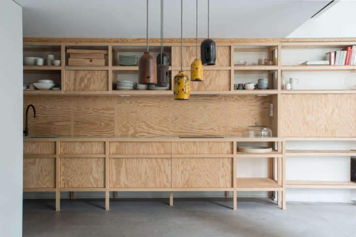 A long wall of plywood shelving that transitions into a fully equipped modern kitchen area.