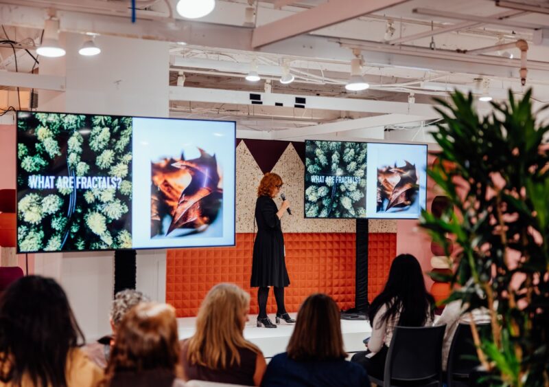 Speaker on stage at NeoCon Talks presenting on fractals to a large audience.