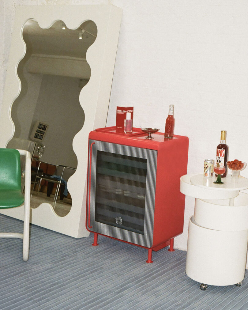 A red mini fridge positioned in a stylish living room next to a wavy mirror and modern furniture.