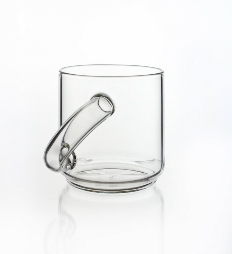 A clear glass mug with a uniquely shaped white handle isolated on a white background.