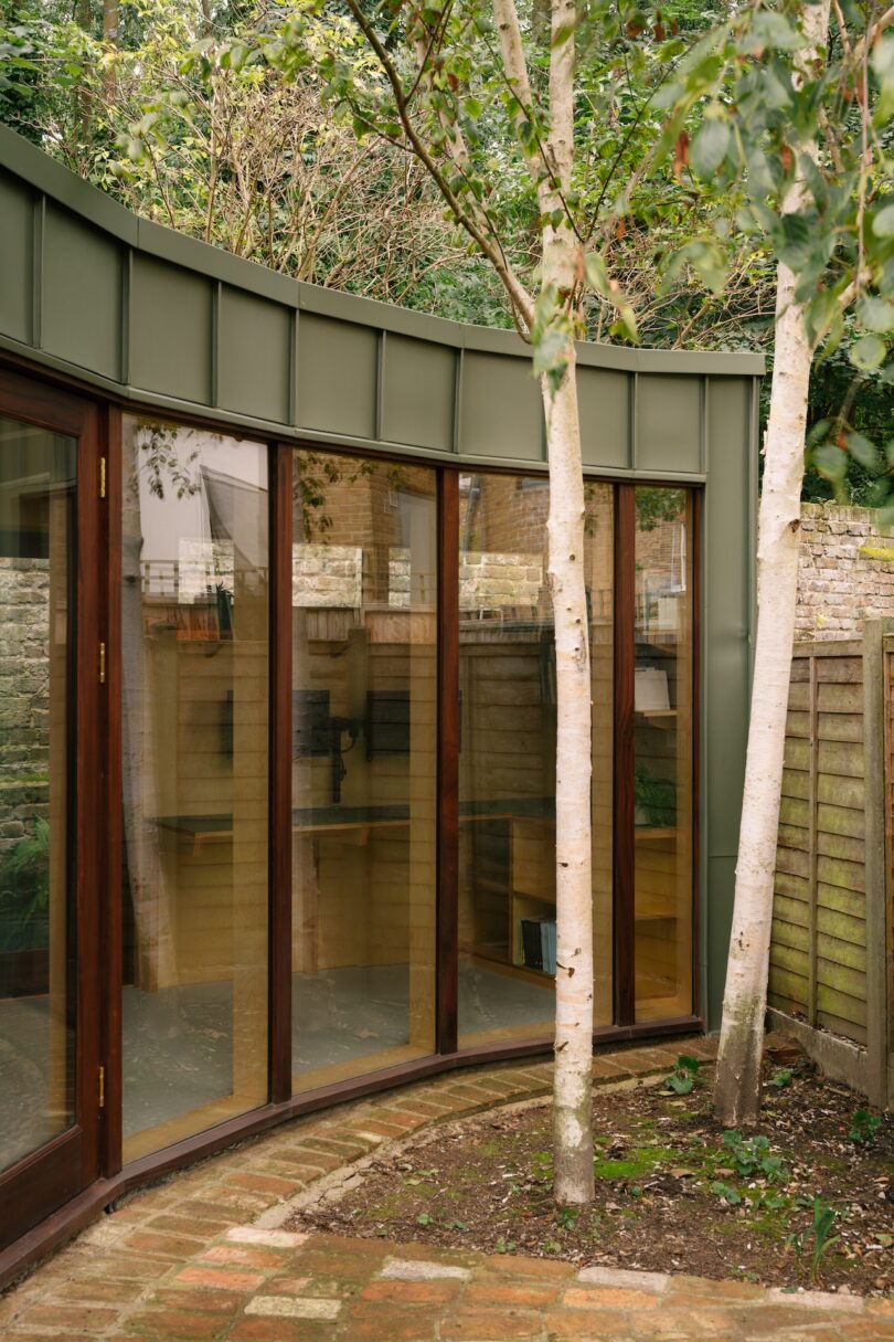 A modern curved building featuring glass windows and green metal cladding surrounded by a wooden fence and trees.