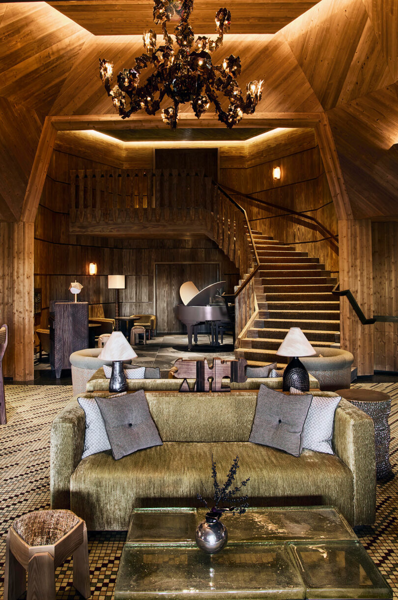 A wood-paneled lounge featuring a gold sofa, a white grand piano, and a sculptural wooden staircase.